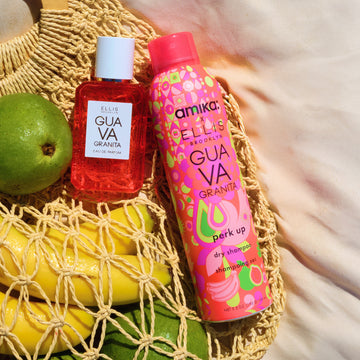 Amika x Ellis Brooklyn GUAVA GRANITA Perk Up Dry Shampoo, GUAVA GRANITA 50ml Eau de Parfum, and a lime lay on top of tan mesh bag filed with bananas and limes against white fabric background
