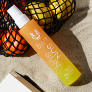 Sun Hair and Body Fragrance Mist 100ml in sand leaning on black mesh bag filled with oranges and lemons, near corner of open book.