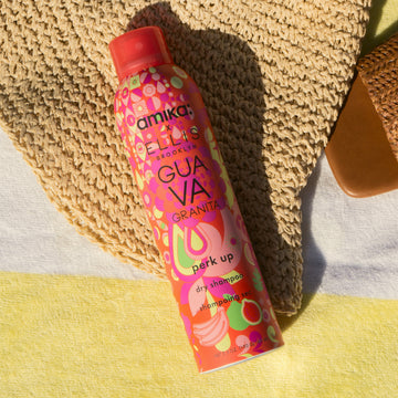Amika x Ellis Brooklyn GUAVA GRANITA Perk Up Dry Shampoo lays on yellow and white striped beach towel and tan, woven hat