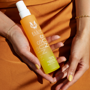Model with yellow nail polish wearing gold bracelets and orange dress holding Sun Hair and Body Fragrance Mist 100ml in one hand on lap.