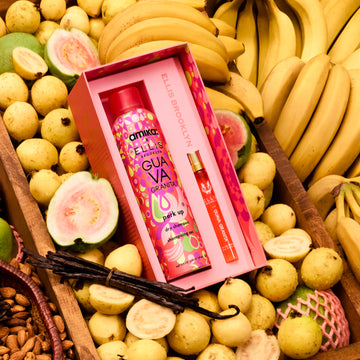 Amika x Ellis Brooklyn GUAVA GRANITA Perk Up Dry Shampoo and GUAVA GRANITA Eau de Parfum 10ml gift set lays in brown wooden bin filled with bananas, whole and sliced guavas, a bundle of vanilla beans, and almonds in a brown basket