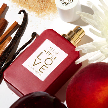 Apple Love 50ml Eau De Parfum with cap removed next to white flower, apple, plum, sandal wood, vanilla beans, and sugar on white background. | english_image