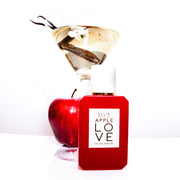 Apple Love 50ml Eau De Parfum on white table in front of apple and martini glass holding white flower, vanilla bean, and water, against white background.