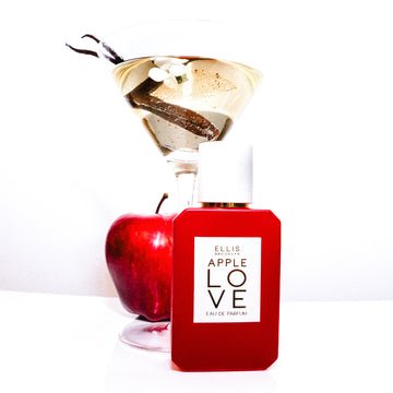 Apple Love 50ml Eau de Parfum sur une table blanche devant une pomme et un verre à martini tenant une fleur blanche, une gousse de vanille et de l'eau, sur fond blanc.