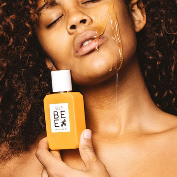 Modèle aux cheveux bruns bouclés tenant Bee 50ml Eau De Parfum contre la mâchoire avec du miel dégoulinant sur la joue, la mâchoire et les lèvres, jusqu'à la main.