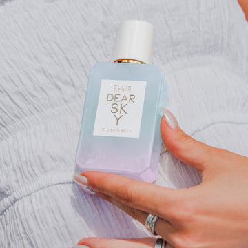 Model with nude nail polish weaing silver rings and white dress holds Dear Sky 50ml Eau De Parfum against stomach.