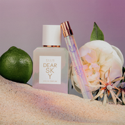 Dear Sky 50ml Eau De Parfum and Dear Sky 10ml Eau De Parfum Travel spray in sand next to Bergamot and flowers, against purple background.