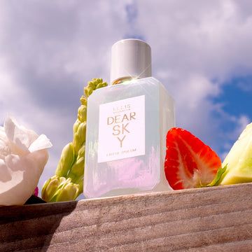 Dear Sky 50ml Eau de Parfum sur un morceau de bois à côté de fraise, melon et fleurs, avec le ciel et les nuages en arrière-plan.