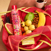 Amika x Ellis Brooklyn GUAVA GRANITA Shampooing sec Perk Up dans un sac amika rose et jaune en mesh avec des goyaves, des gousses de vanille, des amandes, une banane et des oranges à côté d’un pitaya et d’une tasse blanche avec une trace de rouge à lèvres rose remplie de café sur une table brune