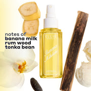 BANANA MILKSHAKE Perfume Mist 240ml  without a cap surrounded by 2 slices of banana, a piece of rum wood, 2 scoops of vanilla ice cream, a tonka bean, and a flower on a white and yellow ombré background with black text that reads 