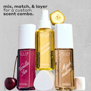 VANILLA SIN Perfume Mist 240ml next to a cherry, BANANA Milkshake Perfume Mist 240ml sitting on top of its cap, MARSHMALLOWS Perfume Mist 240ml with a slice on banana on top of it and a marshmallow next to it, all covered in water droplets on a gray, rough background with black text that reads 