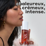Femme aux cheveux noirs et à la frange, portant des boucles d'oreilles créoles en or, un débardeur noir et un pull blanc duveteux, tient le parfum en brume FIRST DATE 90 ml contre son menton avec un texte noir qui dit « chaleureux, crémeux, intense » sur un fond blanc