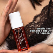 Model with black hair in black lace top and white fuzzy sweater holds FIRST DATE Perfume Mist 90ml between her two palms in front of her chest with white text that reads 
