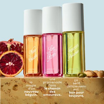 Brume de parfum NEW CRUSH 90ml, Brume de parfum LOVE POM 90ml et Brume de parfum FOREVER PISTACHIO 90ml sur du marbre beige entre des tranches de grenade, d’orange sanguine et des pistaches, sur un fond bleu clair avec un texte blanc qui dit « la magie d’un nouveau béguin. Une promenade dans le chemin des amoureux. Ca sent délicieusem bon pour toujours. »