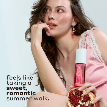 Woman with brown hair wearing pink silk and white lace tank top and pink earrings holds LOVE POM Perfume Mist 90ml on half of a pomegranate and places hand against her lips against a gray background with black text that reads 