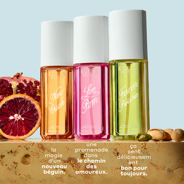 Brume de parfum NEW CRUSH 90ml, Brume de parfum LOVE POM 90ml et Brume de parfum FOREVER PISTACHIO 90ml sur du marbre beige entre des tranches de grenade, d’orange sanguine et des pistaches, sur un fond bleu clair avec un texte blanc qui dit « la magie d’un nouveau béguin. Une promenade dans le chemin des amoureux. ça sent délicieusement bon pour toujours. »