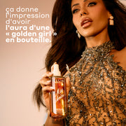 Un flacon de parfum Miss Amber Perfume Mist avec le bouchon enlevé, tenu par un mannequin aux cheveux foncés portant un haut orné de bijoux, avec du texte à gauche qui dit « ca donne l'impression d'avoir l'aura d'une golden girl en bouteille ».