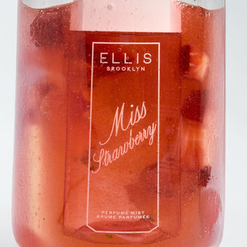 Brume parfumée MISS STRAWBERRY 90 ml dans un verre transparent rempli de jus rouge et de fraises coupées sur fond blanc