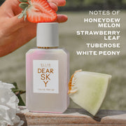 DEAR SKY Eau de Parfum 50ml between slice of honeydew melon and white flower with a hand placing a sliced strawberry on the cap sit on wood against a background of grass with white text that reads 
