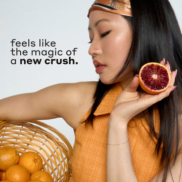 Woman with black hair wearing an orange and blue headband and tank top holds tan basket filled with oranges and a sliced blood orange against white background with black text that reads 
