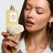 Une femme aux cheveux noirs, vêtue d'une chemise blanche et portant des créoles dorées, tient un cornet de glace à la vanille surmonté du flacon d'eau de parfum VANILLA SALT (50 ml, sans capuchon), le tout sur fond gris.