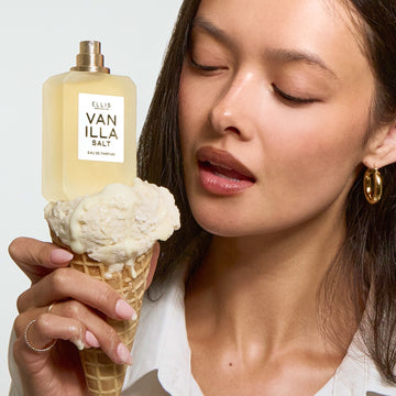 A woman with black hair wearing a white shirt and gold hoop earrings holds a vanilla ice cream cone with VANILLA SALT Eau de Parfum 50ml without cap on top all on a gray background.