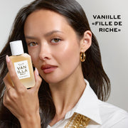 Une femme aux cheveux noirs, vêtue d'une chemise blanche et portant un collier ainsi que des créoles en or, tient un flacon d'eau de parfum VANILLA SALT (50 ml) près de son visage, sur un fond gris où figure le texte noir « Vanille « Fille de Riche » ».