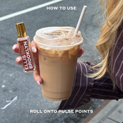 Model with nude nail polish and blonde hair wearing a brown and white stripped long sleeve button-down shirt holding and iced coffee and Vanilla Santo Fragrance Oil 15ml in one hand on city street between white text that reads 