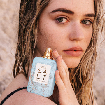Woman with brown, wet hair wearing a black bikini holds SALT Eau de Parfum 50ml without cap on her shoulder all covered in sand against tan beach background