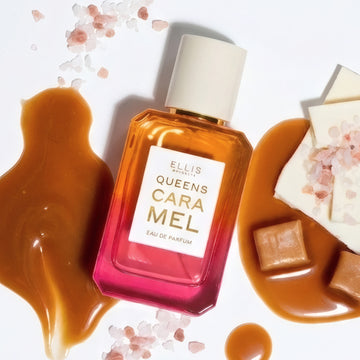 QUEENS CARAMEL Eau de Parfum 50ml lays next to three puddles of caramel, two piles of pink salt, two caramel candies, and pieces of white chocolate with a pile of pink salt on top on a white background