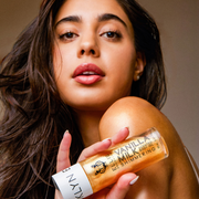 Model in tank top with brown hair and white nail polish holding Vanilla Milk Shimmering Scented Body Oil near shoulder with brown background.