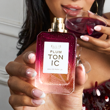 Woman with brown hair wearing maroon sequined tank top holds PLUM TONIC Eau de Parfum 50ml without cap covered in water droplets in one hand and a glass coupe with a plum slice filled with purple liquid against her lips in the other against a white wall