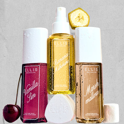 VANILLA SIN Perfume Mist 240ml next to a cherry, BANANA Milkshake Perfume Mist 240ml sitting on top of it's cap, MARSHMALLOWS Perfume Mist 240ml with a slice on banana on top of it and a marshmallow next to it, all covered in water droplets on a gray, rough background 