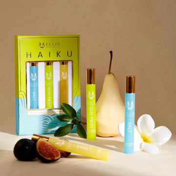 Green, blue, and gold Haiku Fragrance Rollerball Trio Box showing Salt 10ml Fragrance Rollerball, Sun Fruit 10ml Fragrance Rollerball, and Sweet 10ml Fragrance Rollerball, on white background on beige backdrop with Salt 10ml Fragrance Rollerball, Sun Fruit 10ml Fragrance Rollerball, Sweet 10ml Fragrance Rollerball, pear, white flower, figs, and green leaves.