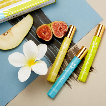 Corner of green, blue, and gold Haiku Fragrance Rollerball Trio Box showing Sweet 10ml Fragrance Rollerball, on beige and blue backdrop with black brush stroke, next to Salt 10ml Fragrance Rollerball, Sun Fruit 10ml Fragrance Rollerball, Sweet 10ml Fragrance Rollerball, pear, white flower, and figs.