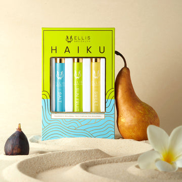 Green, blue, and gold Haiku Fragrance Rollerball Trio Box showing Salt 10ml Fragrance Rollerball, Sun Fruit 10ml Fragrance Rollerball, and Sweet 10ml Fragrance Rollerball, in sand next to pear, fig, and white flower, against beige background.