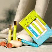 Green, blue, and gold Haiku Fragrance Rollerball Trio Box showing Salt 10ml Fragrance Rollerball, Sun Fruit 10ml Fragrance Rollerball, and Sweet 10ml Fragrance Rollerball, next to Salt 10ml Fragrance Rollerball, Sun Fruit 10ml Fragrance Rollerball, Sweet 10ml Fragrance Rollerball, and figs resting on stones, against beige fabric background.