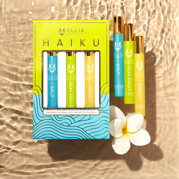 Boîte trio de flacons roll-on parfumés Haiku vert, bleu et or, comprenant le roll-on parfum Salt 10ml, le roll-on parfum Sun Fruit 10ml et le roll-on parfum Sweet 10ml, sur un fond beige effet eau avec les roll-ons Salt 10ml, Sun Fruit 10ml, Sweet 10ml et une fleur blanche.