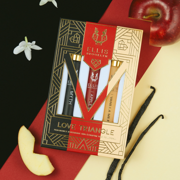Black, red, beige and gold Love Triangle Fragrance Rollerball Trio Box showing Myth 10ml Fragrance Rollerball, Apple Love 10ml Fragrance Rollerball, and Vanilla Milk 10ml Fragrance Rollerball, on black, red, and beige background with white flowers, apple, and vanilla beans.