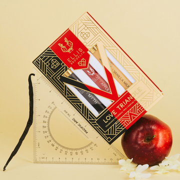 Black, red, beige and gold Love Triangle Fragrance Rollerball Trio Box showing Myth 10ml Fragrance Rollerball, Apple Love 10ml Fragrance Rollerball, and Vanilla Milk 10ml Fragrance Rollerball, leaning on measuring tool and apple with vanilla bean and white flowers, on light yellow backdrop.