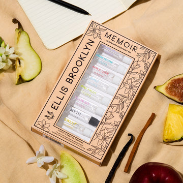Boîte de découverte de parfums Memoir avec un design floral beige et noir montrant les flacons de 1,5 ml Dear Sky, Miami Nectar, Apple Love, Salt, Sun Fruit, Florist, Myth, Sweet et Vanilla Milk, posés sur un tissu marron clair avec figue, ananas, pomme, bois de santal, gousse de vanille, poire, fleurs blanches et un carnet avec stylo.