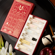 Une boîte rouge Ellis Brooklyn Mystery Fragrance Discovery Set ouverte montrant à l'intérieur : Flacon de 1,5 ml Apple Love, Flacon de 1,5 ml Florist, Flacon de 1,5 ml Salt, Flacon de 1,5 ml Myth, Flacon de 1,5 ml Vanilla Milk et Flacon de 1,5 ml Sweet sur un disque vinyle noir avec des gousses de vanille, des guimauves, une pomme, du bois de santal et des fleurs blanches, sur une table en bois.