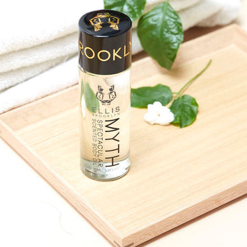 Huile corporelle parfumée Myth Spectacular 95 ml et fleur blanche sur une planche en bois, à côté de serviettes blanches pliées et de feuilles vertes, sur une table blanche.