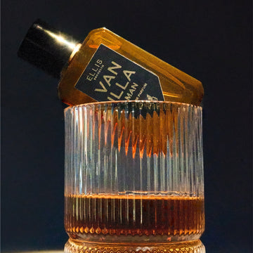 Vanilla Man 50ml Eau De Parfum sitting on rim of glass of whiskey, on black background. | english_image
