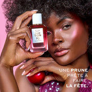 Une femme aux cheveux bruns portant un débardeur à sequins violet et du surligneur rose tient PLUM TONIC Eau de Parfum 50 ml contre sa tête d'une main et une prune de l'autre sur un fond rose avec un texte blanc qui dit « Une prune prête à faire la fête. »