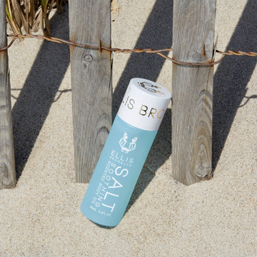 Huile corporelle parfumée apaisante Salt 95ml dans le sable, appuyée contre une clôture en bois et métal avec de l'herbe de plage en arrière-plan.
