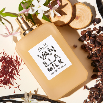Lait à la vanille 50ml Eau de Parfum sans bouchon sur fond blanc cassé entouré de bois de cèdre, de fleurs blanches, de gousses de vanille, de safran et de coques de cacao.