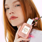 Woman with red hair wearing a pink shirt holds VANILLA RICE Eau de Parfum 50ml and pink cherry blossoms against her shoulder on a pink background
