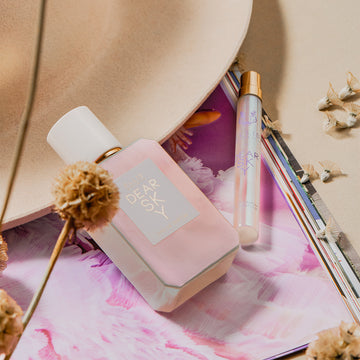 Le parfum Dear Sky 50ml Eau De Parfum et le spray voyage Dear Sky 10ml Eau De Parfum reposant sur une image de magazine violet et blanc et un chapeau beige avec des fleurs séchées, sur un fond beige.