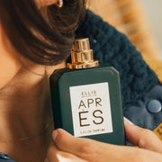 Model with brown hair and nude nail polish wearing gold necklace, gold diamond earring and blue fleece holding Apres 50ml Eau De Parfum with cap removed near neck, against tan background.